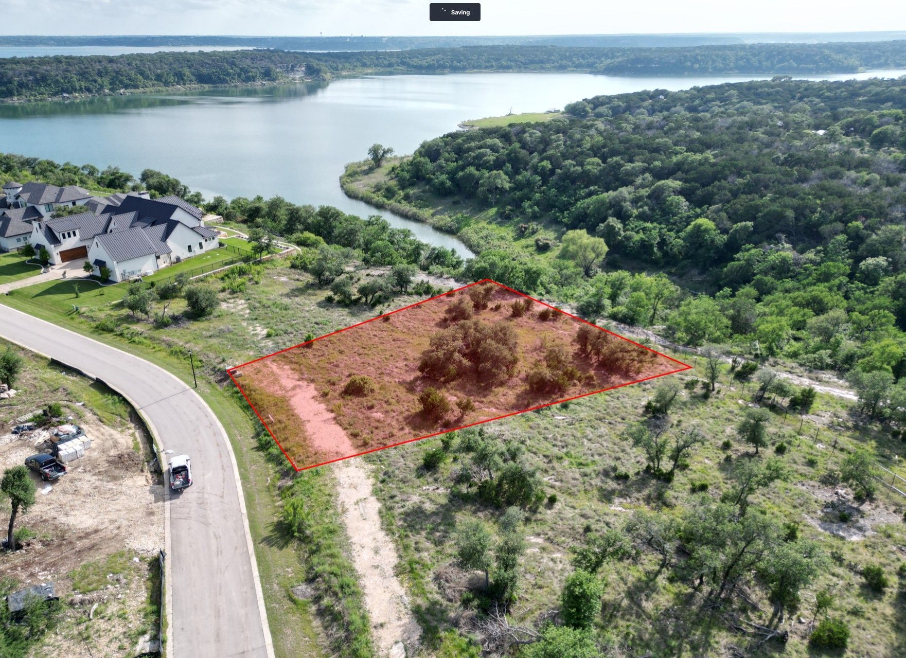 Aerial view of a lake, wooded hills, and a marked empty lot beside a winding road