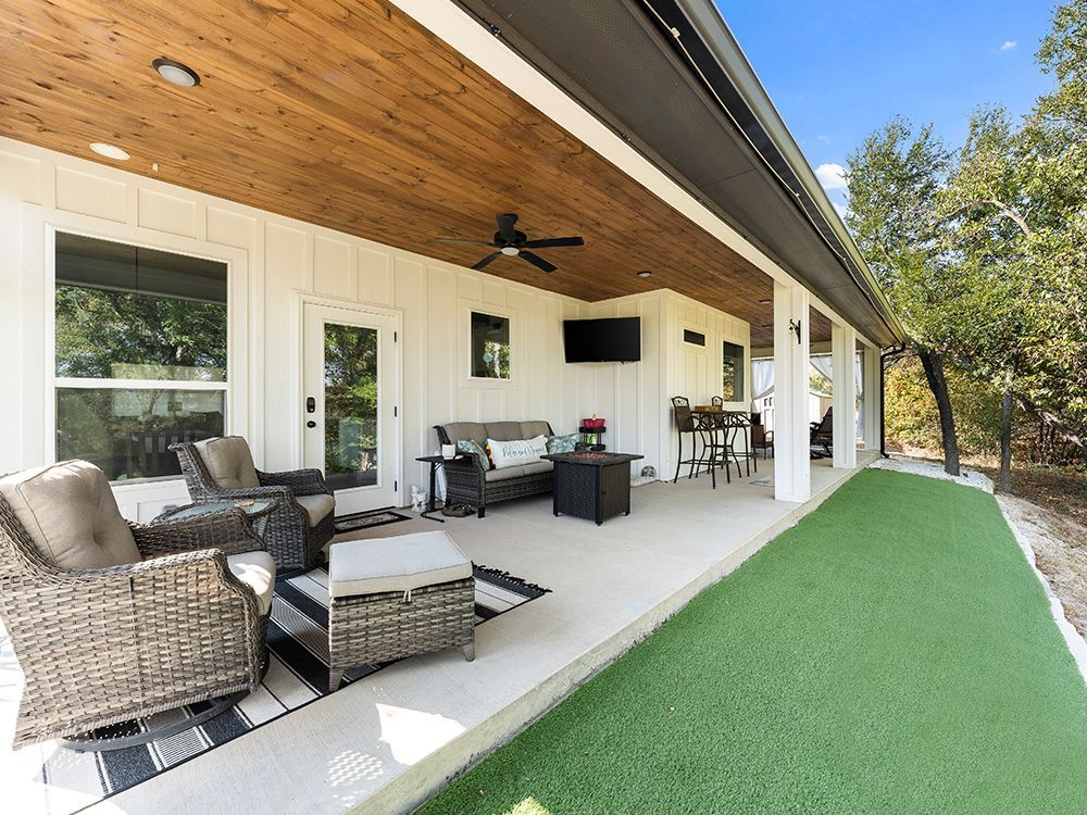 Covered outdoor patio with seating, television, and artificial grass. White walls, wood ceiling, and surrounding trees.