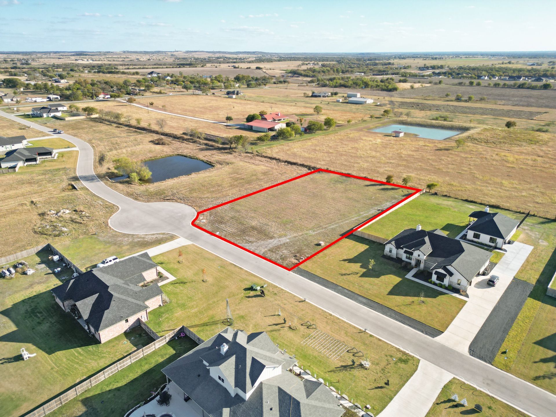 Aerial view of vacant residential lot outlined in red, surrounded by houses and a rural landscape.
