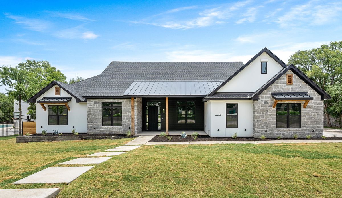 Modern, single-story house with gray stone and white accents, black windows, and a green lawn under a blue sky.