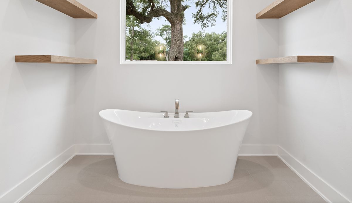 White freestanding bathtub in a minimalist bathroom with wood shelves and a window overlooking a tree.