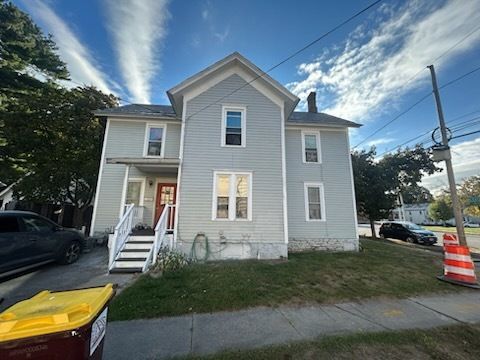 Two-story, light gray house with white trim, green lawn, blue sky. Yellow trash bin in front.