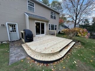 Backyard deck with curved edges, stairs, and a grill. Attached to a beige house, lawn in front.
