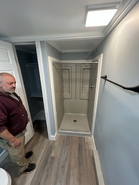 Bathroom with shower stall and man observing. Light wood flooring, blue-gray walls, and white trim.