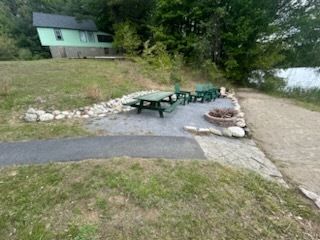 Green picnic tables and fire pit on a gravel patio beside a paved path and lake, with a green house in background.