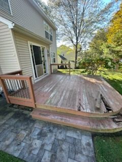 Exterior view of a weathered wooden deck attached to a two-story house, with steps leading down to a stone patio.