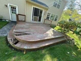 Damaged wooden deck with broken boards and steps, attached to a light-colored house. Green grass surrounds it.