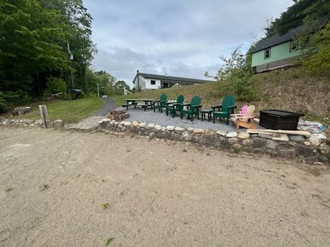 Outdoor seating area with Adirondack chairs, fire pit, and picnic table on gravel. Green building in the background.