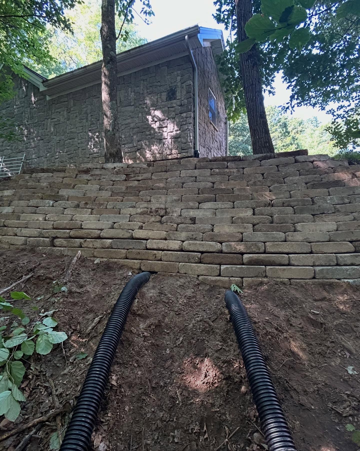 Retaining wall built on a hillside, leading to a stone building. Two black drainage pipes visible at the base of the wall.