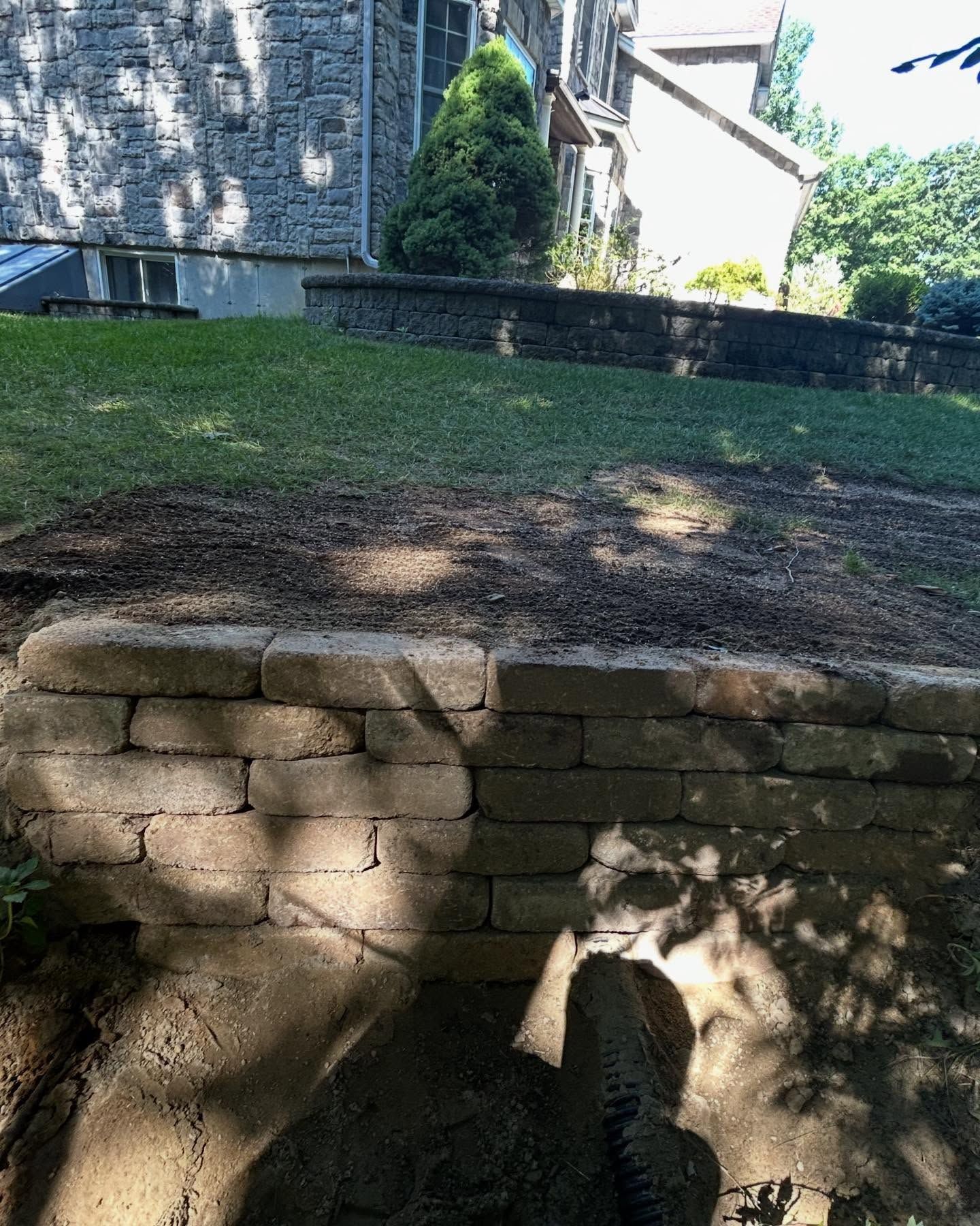 Stone retaining wall supporting a grassy hill, with a building in the background.