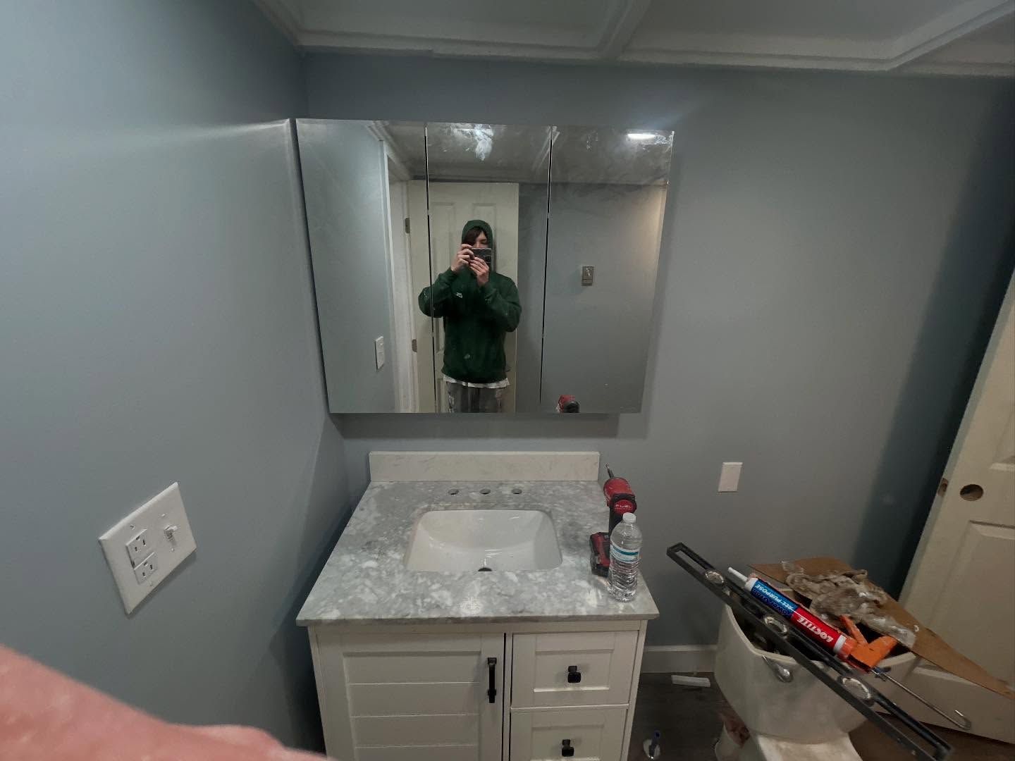 Bathroom with a person taking a photo in the mirror. Gray walls, white vanity, and a partially open door.
