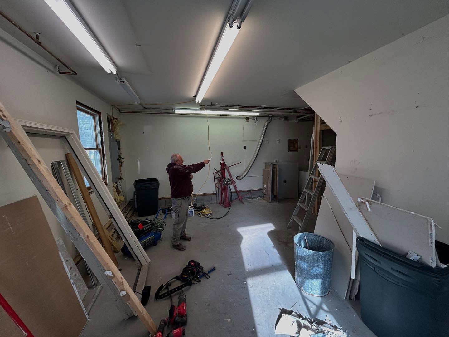 Man in work clothes gesturing in a room under construction, with tools, debris, and overhead lights.