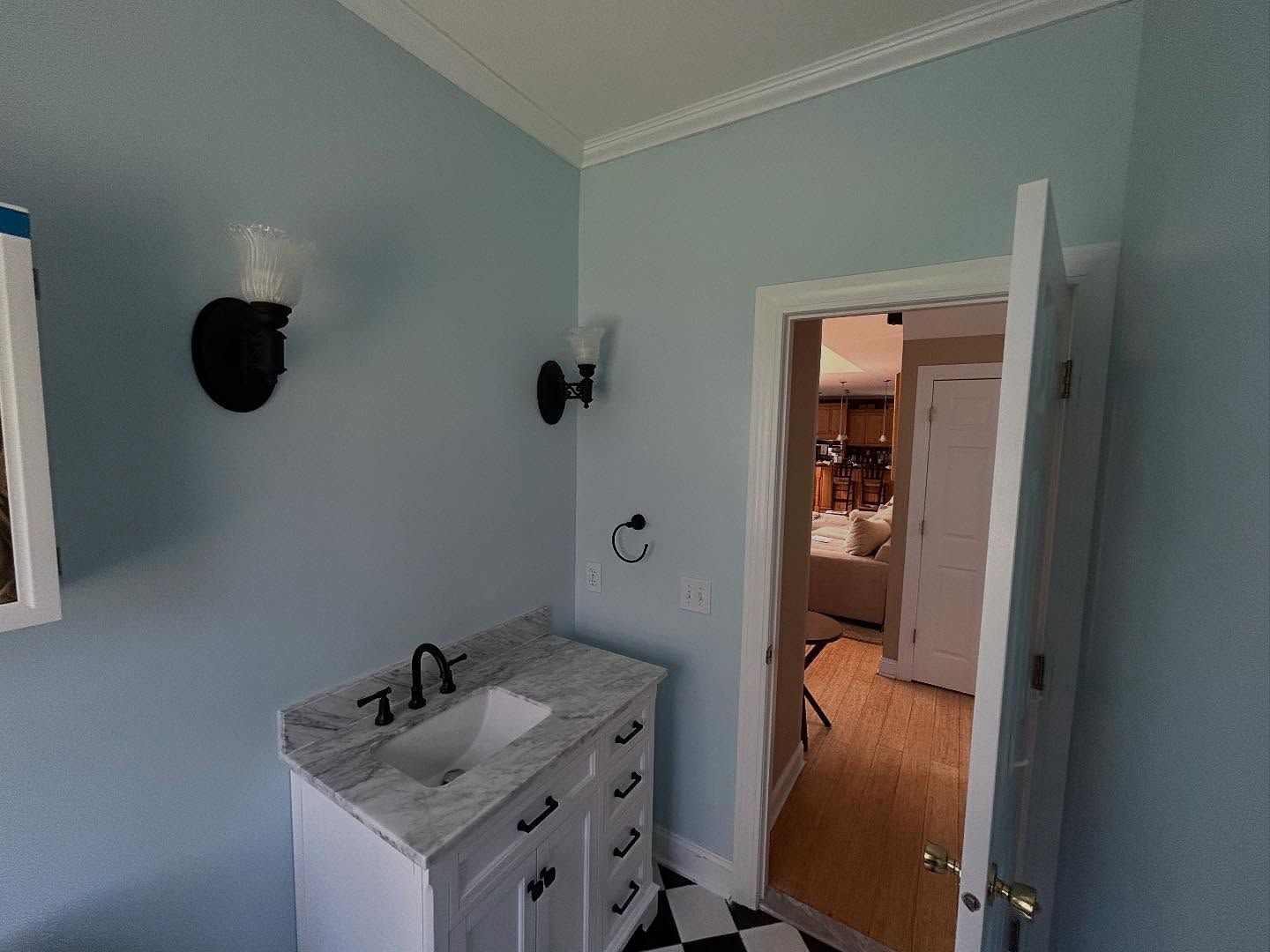 Bathroom with a vanity, black fixtures, and a doorway leading to a room. Light blue walls, black and white floor.