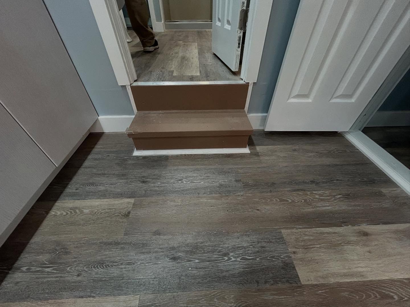 A hallway with wood-look flooring and a small step up to a door. A person is visible in the doorway.