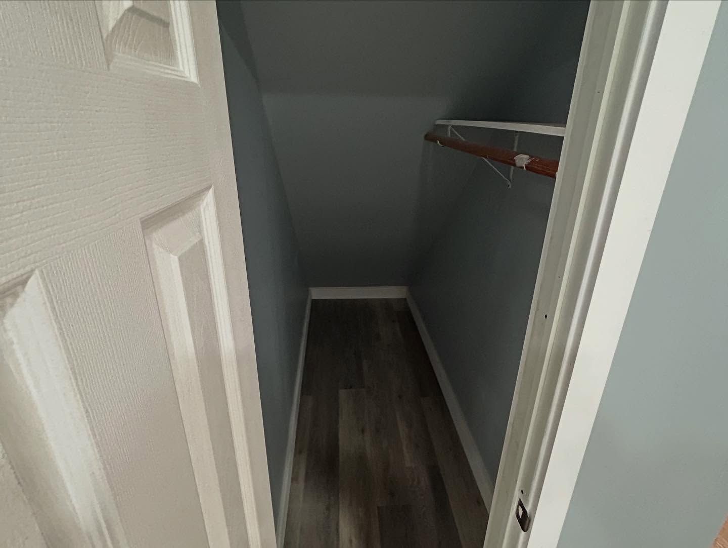 Empty, walk-in closet with angled walls, wooden shelf and flooring, and a white door.