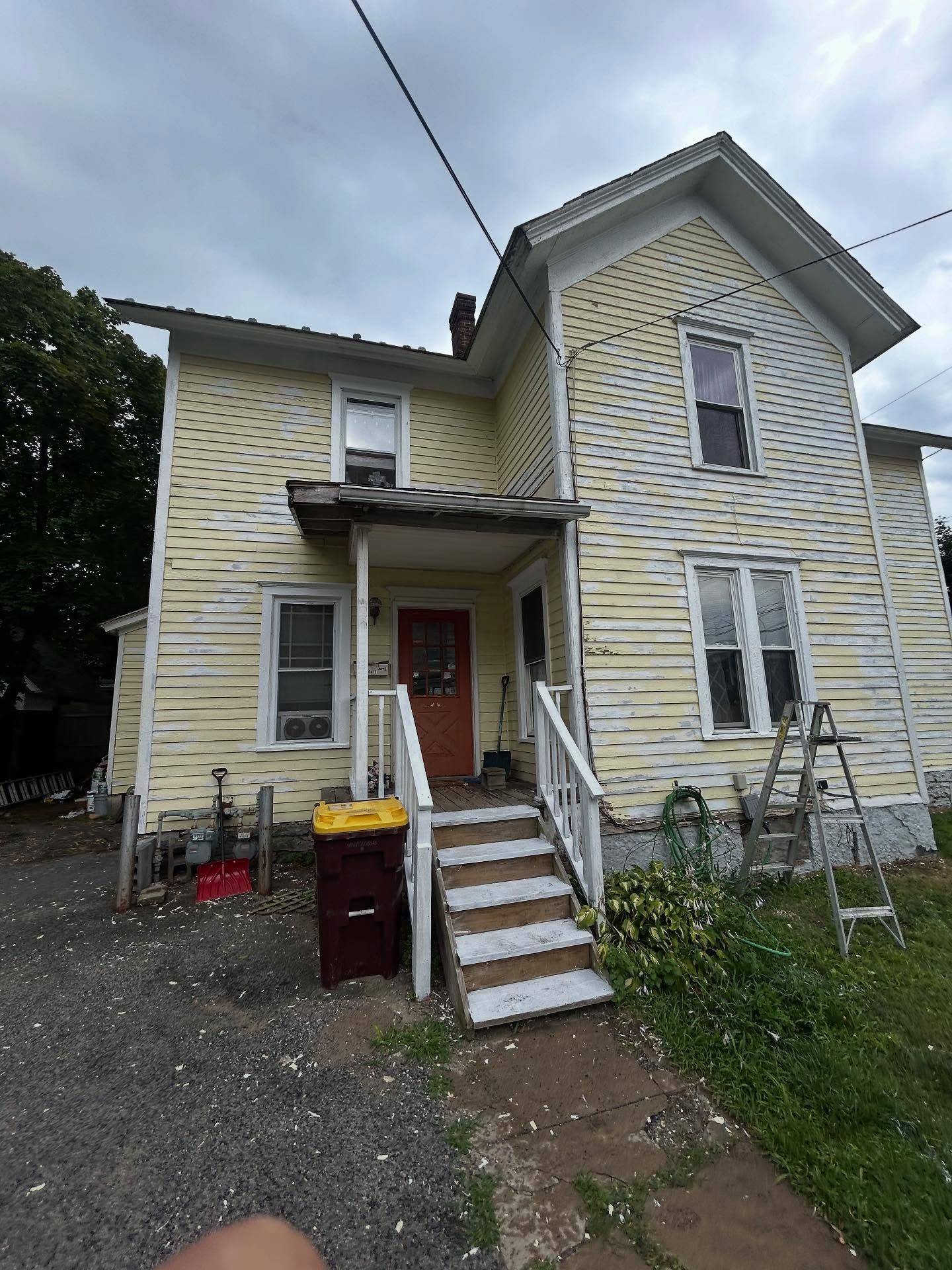 Yellow two-story house with peeling paint, front steps, and a garbage can in front. Overcast sky.