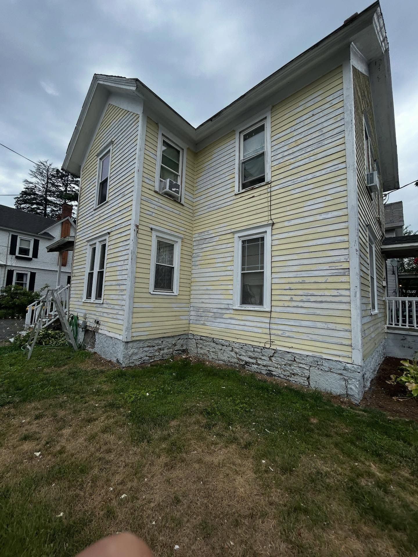 Two-story, yellow clapboard house with peeling paint, cloudy sky background.