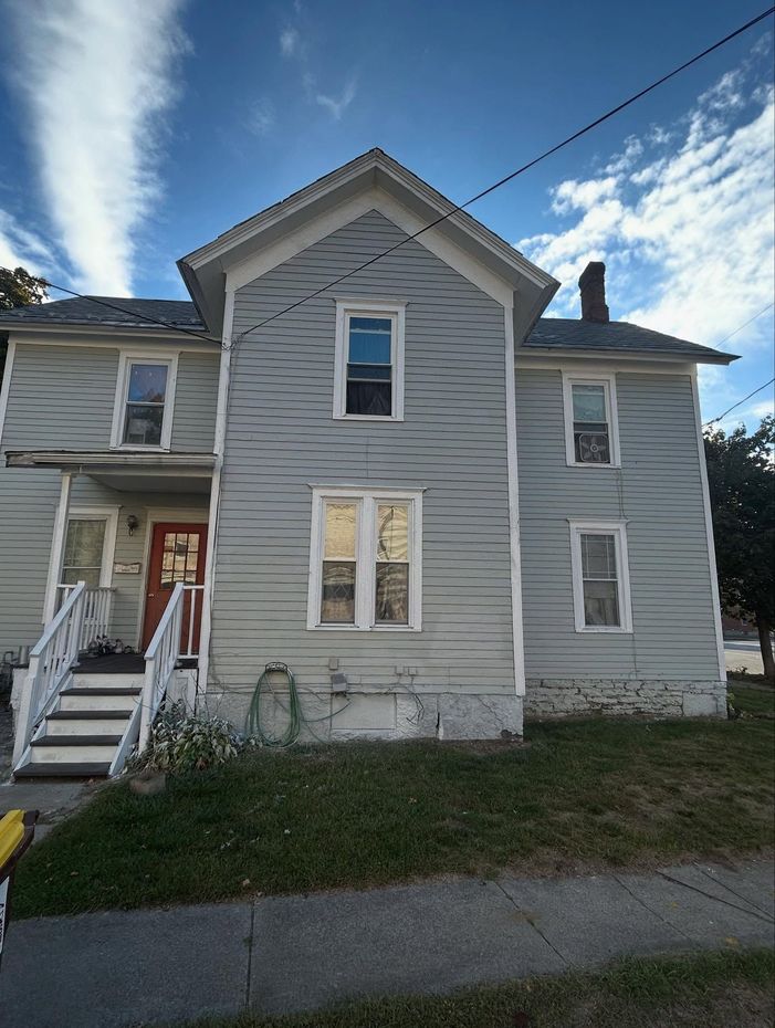 Two-story gray house with white trim. Red front door, stairs leading to the porch. Cloudy blue sky.