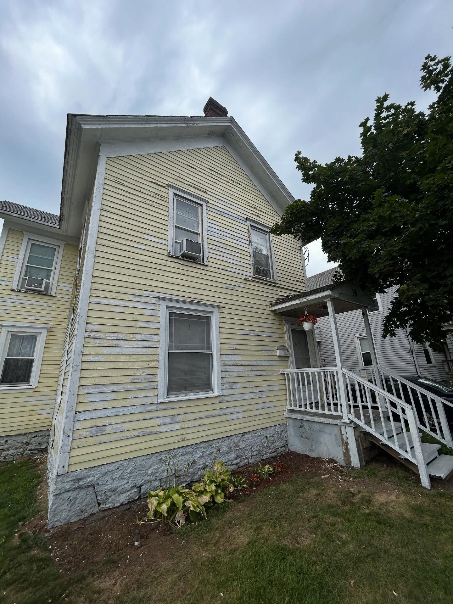 Yellow house with white trim, chimney, and front porch with stairs. Overcast sky.