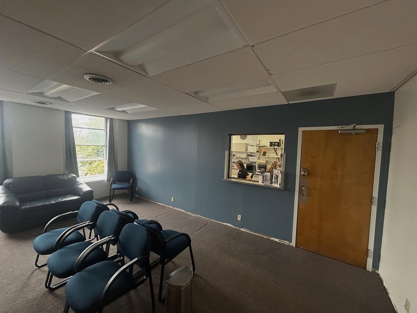 Waiting room with blue chairs, sofa, and window to a counter. Blue wall.