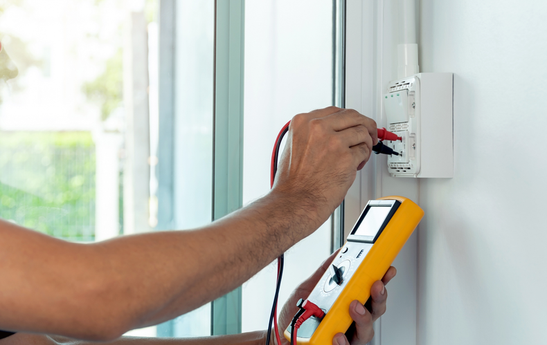 Person using a multimeter to test a white electrical outlet on a white wall.