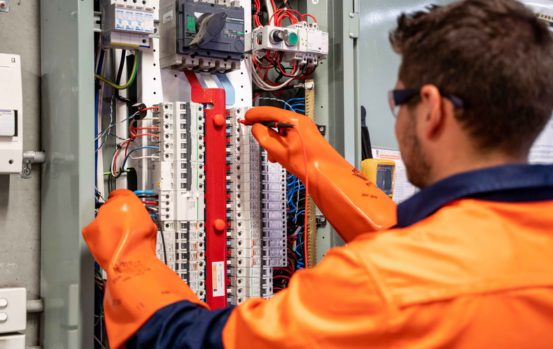 Electrician working on electrical panel, wearing safety glasses and orange gloves.