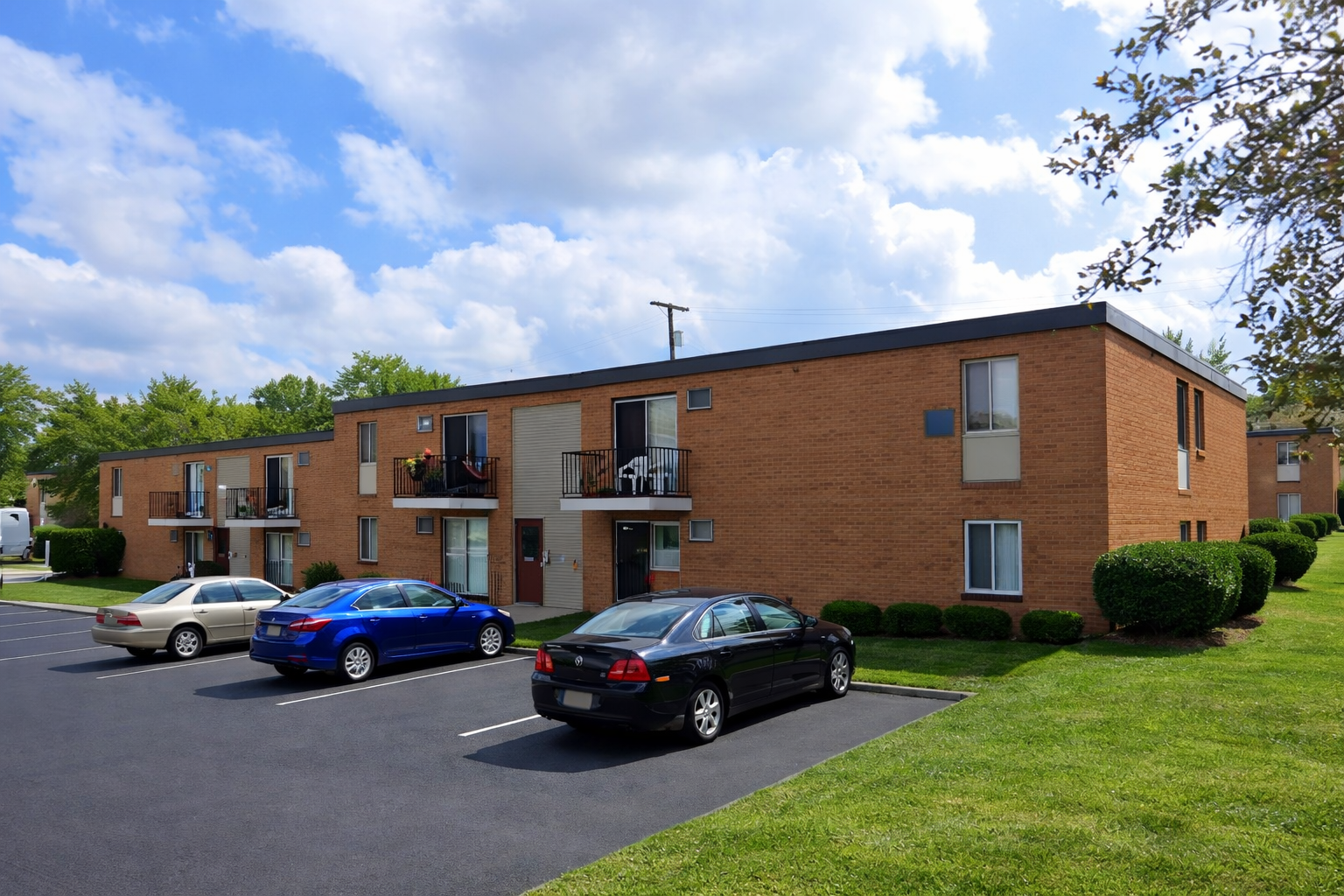 A two-story red brick apartment building with balconies and parked cars in the foreground under a blue sky with clouds.