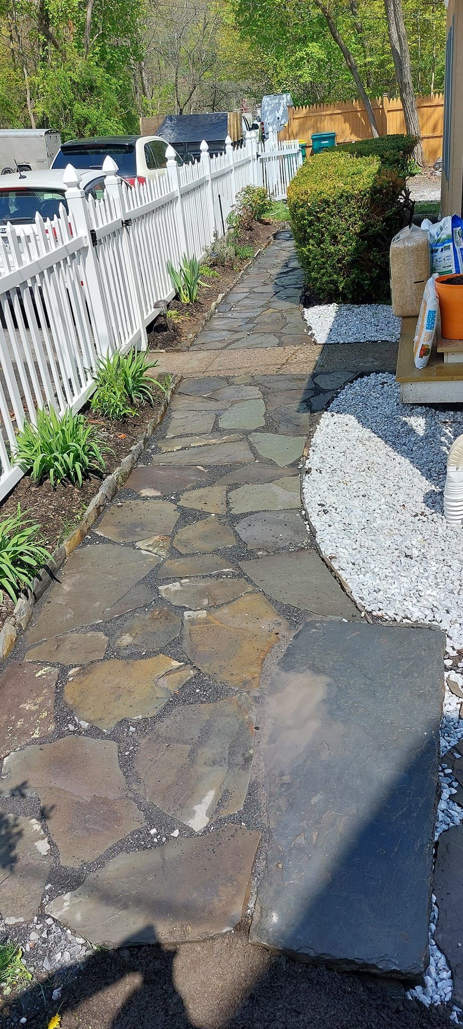 Stone pathway next to a white picket fence, leading to a house entrance. Green foliage and trees in the background.