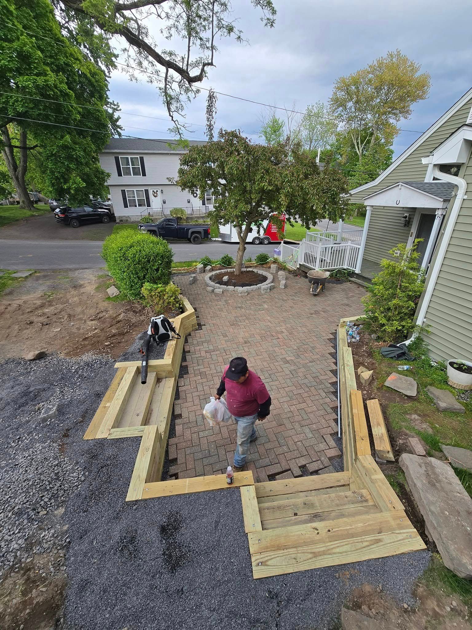 Man walking on a stone path towards a house, carrying bags. Wooden stairs, a gravel base, and a tree are also visible.