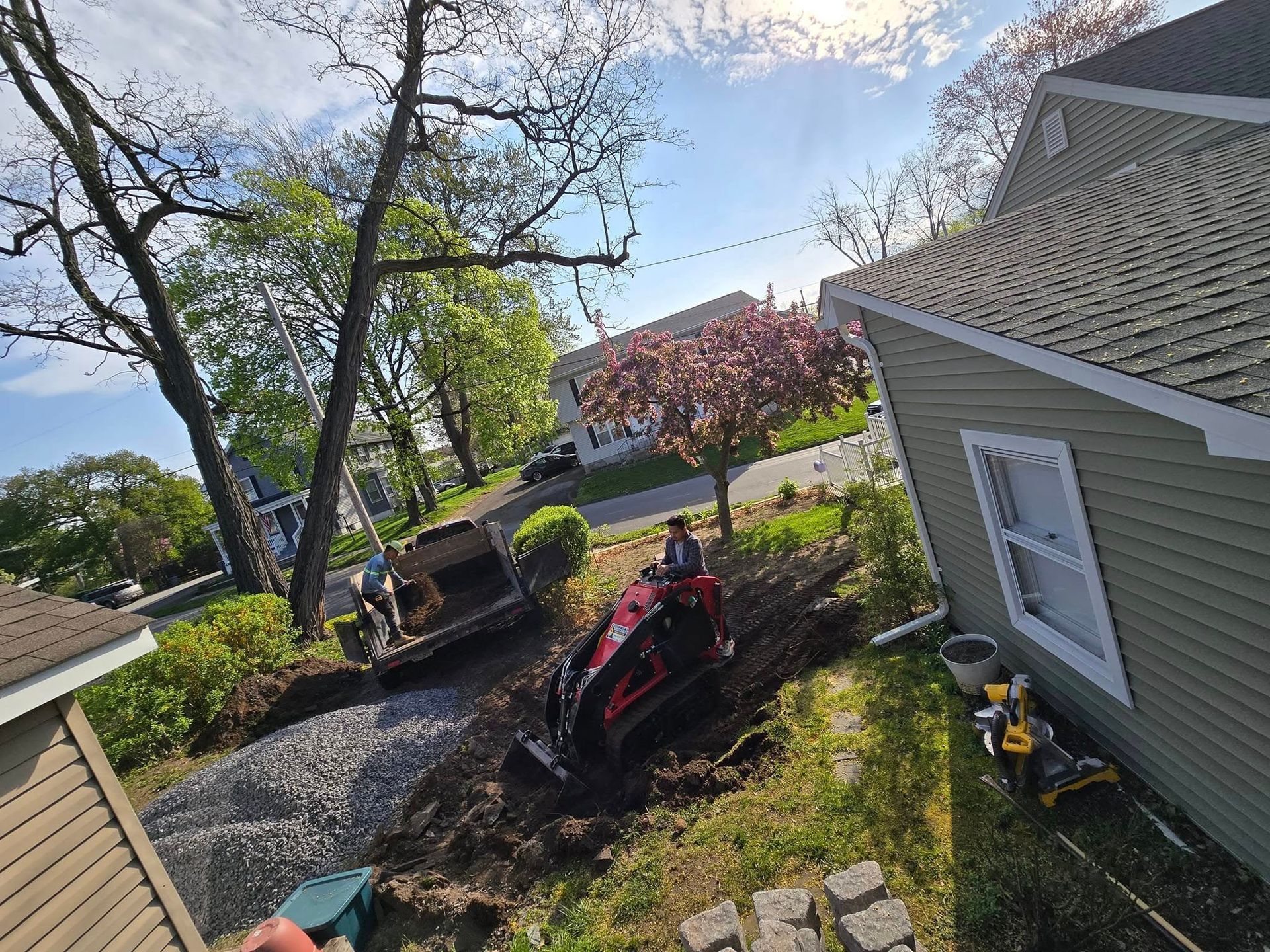Construction workers using machinery to landscape yard next to a green house, sunny day.