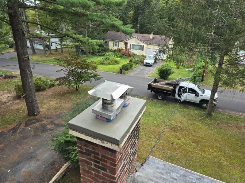Brick chimney with a metal cap. A white truck with a load of wood is parked nearby a house.