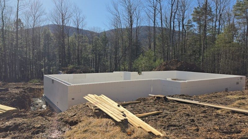 Concrete foundation of a building under construction, surrounded by dirt, wood, and trees.
