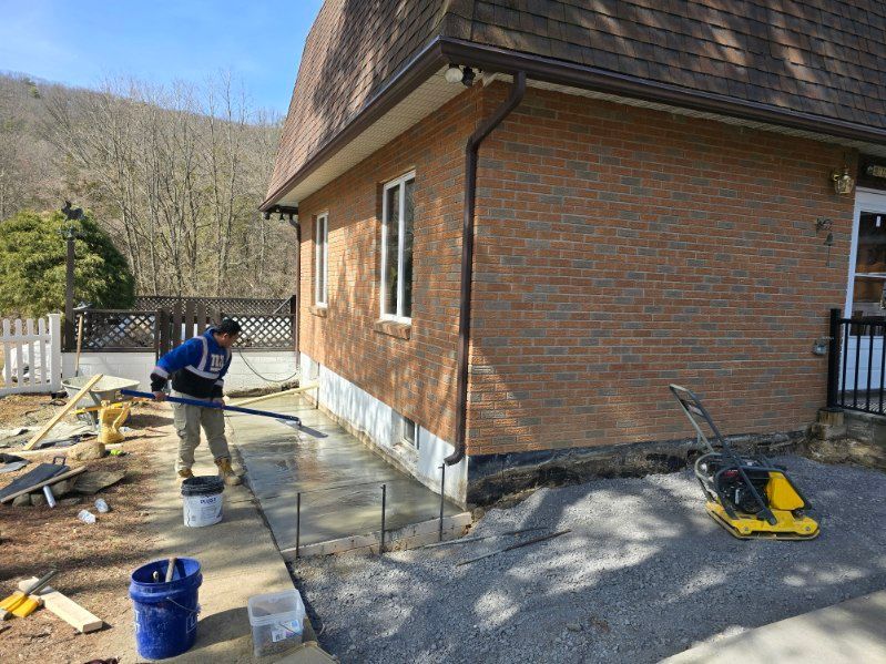 Man smoothing wet concrete sidewalk next to brick building. Gravel and construction materials visible.
