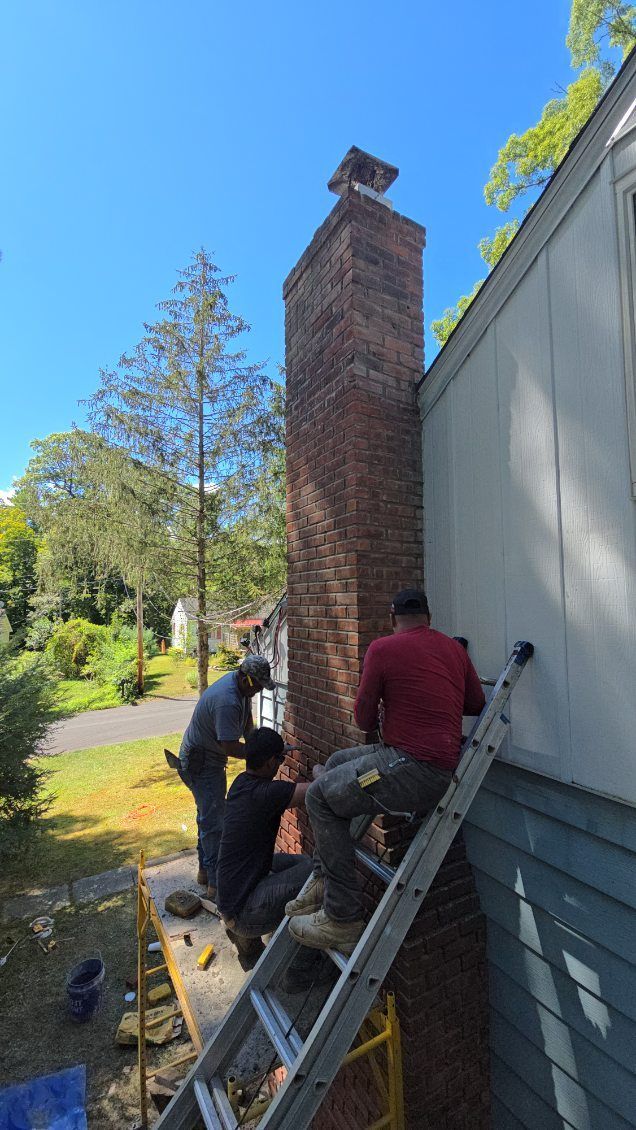 Three workers repair a brick chimney on a house, using a ladder on a sunny day.