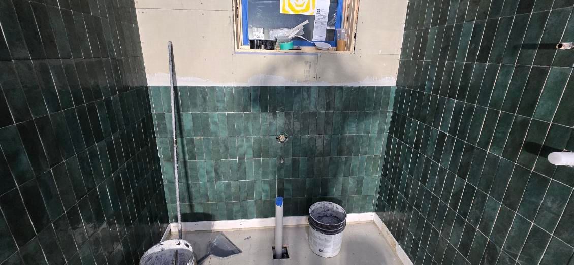 A bathroom with green tile walls and a window. A mop and bucket sit on the floor.