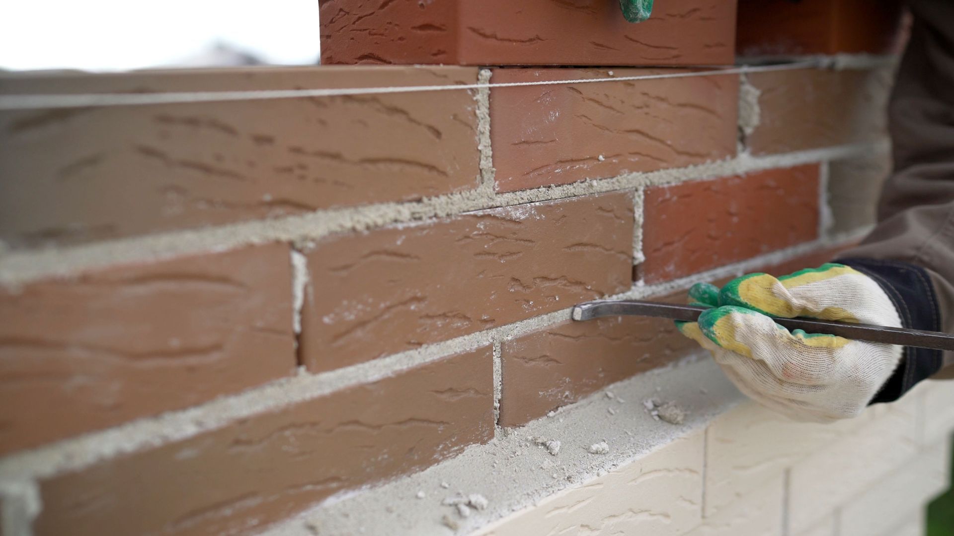 A gloved hand uses a tool to smooth mortar between red and brown bricks on a building.