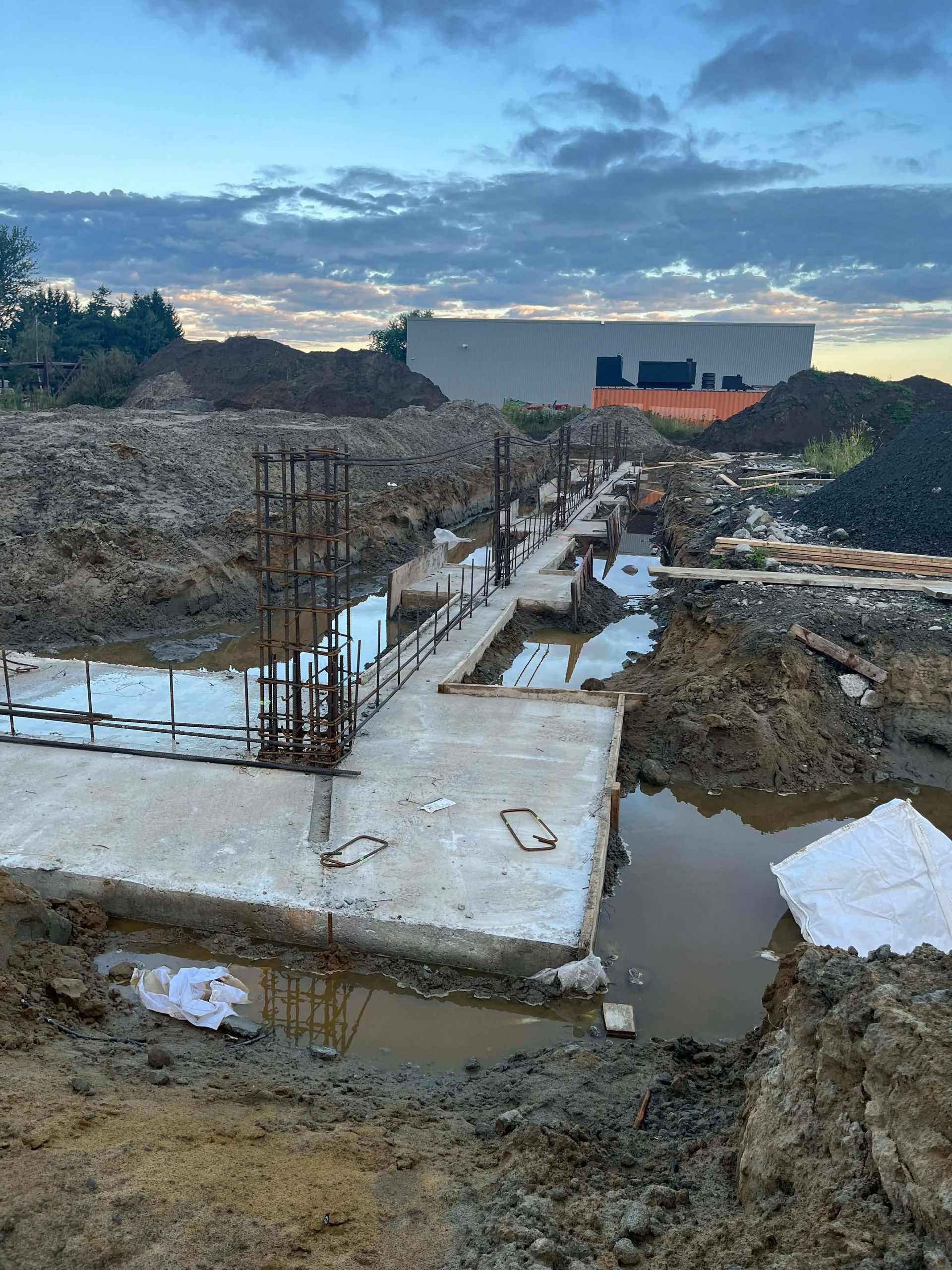 Fondation en béton en construction à côté d'une excavation boueuse au crépuscule, avec des barres d'armature et un bâtiment au loin.
