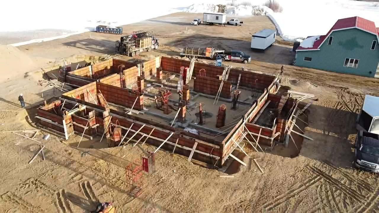 Vue aérienne des fondations d'une maison en construction dans une zone sablonneuse à proximité d'autres habitations.