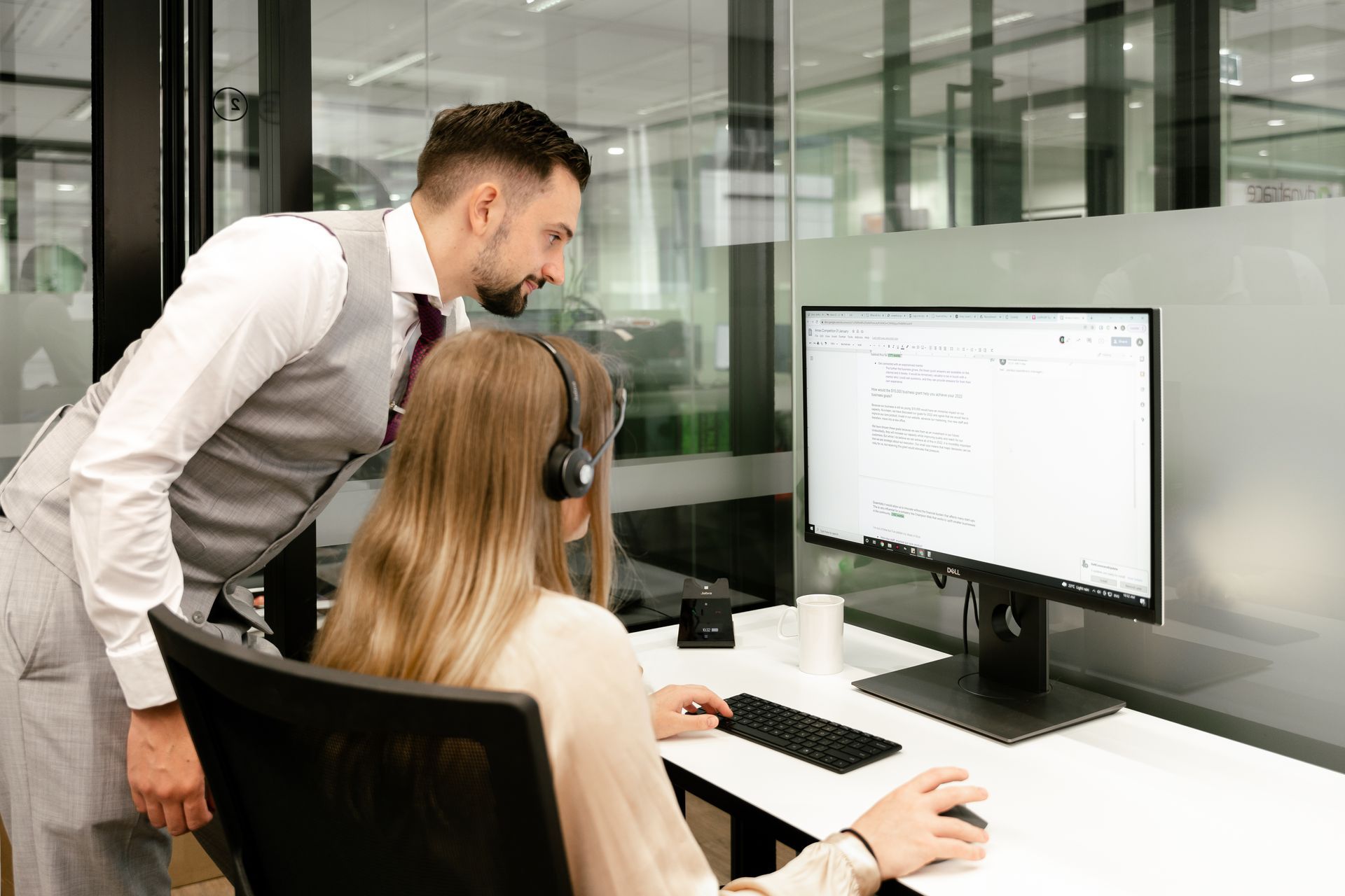 A man is standing next to a woman sitting in front of a computer.