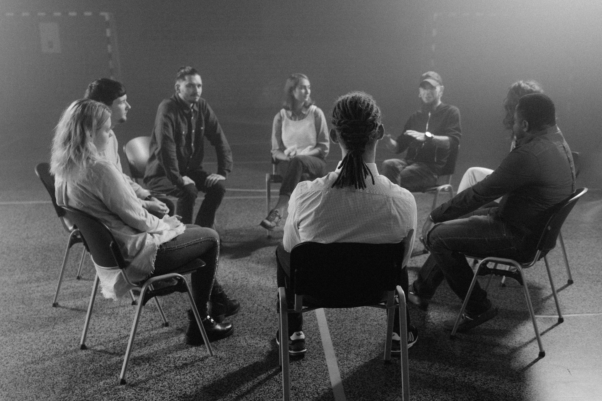Group therapy session in a dimly lit room; diverse people sitting in a circle, facing each other, engaged in a discussion.
