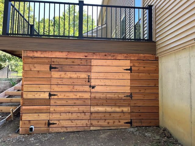 Wooden storage doors built under a deck, with black hardware. House exterior, green trees in background.
