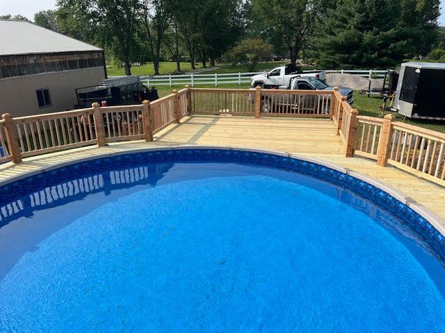 Wooden deck surrounds a blue, in-ground pool. A barn and trees are in the background.