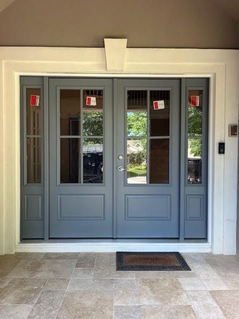 Gray double doors with glass panels, flanked by sidelights, set in a white frame, with a welcome mat on a tiled porch.
