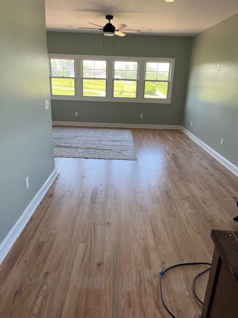 Light wood floors, pale green walls, and large windows in a spacious room with a neutral rug and ceiling fan.