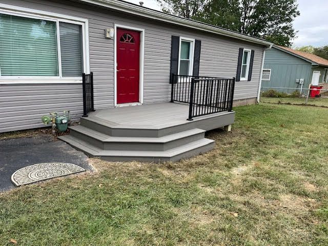 Gray house with a red door, a deck, and steps leading to the entrance. Black railings surround the deck.