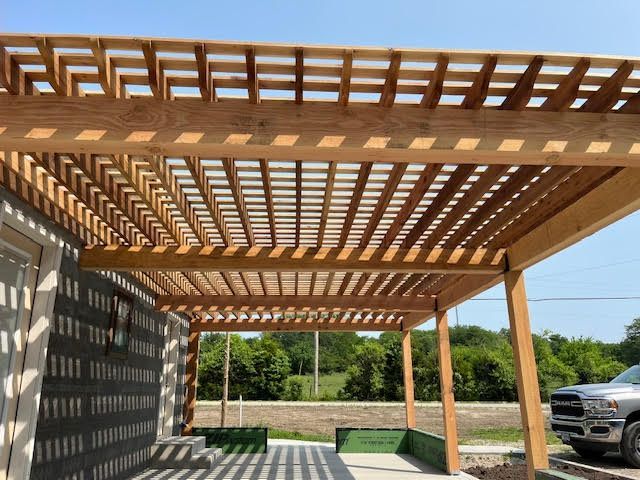 Wooden pergola attached to a building, providing shade. Sunlight casts shadows on the ground.