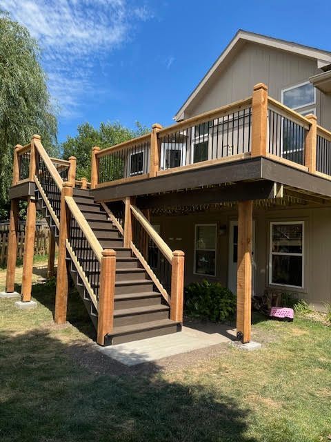 A two-story deck with stairs leading down to a concrete patio in a backyard setting; brown wood and black railing.