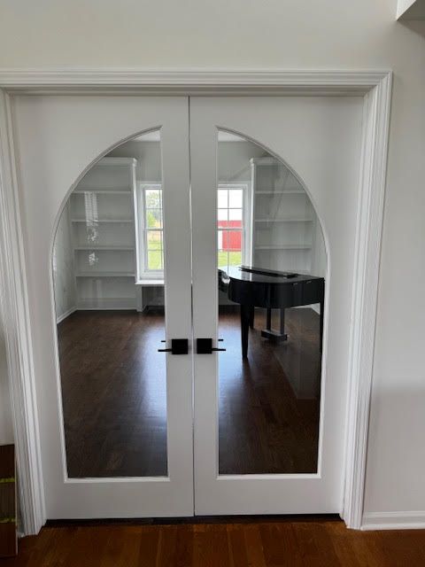 White double doors with arched tops, glass panes, leading to a room with a piano and bookshelves.