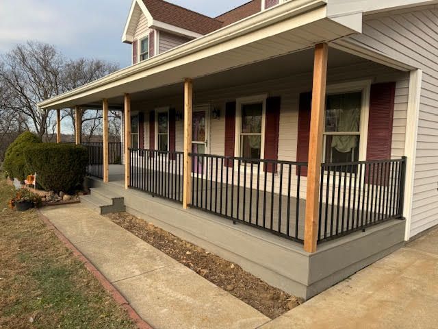 A porch with black railings, wooden posts, and a gray base, in front of a house with red shutters.