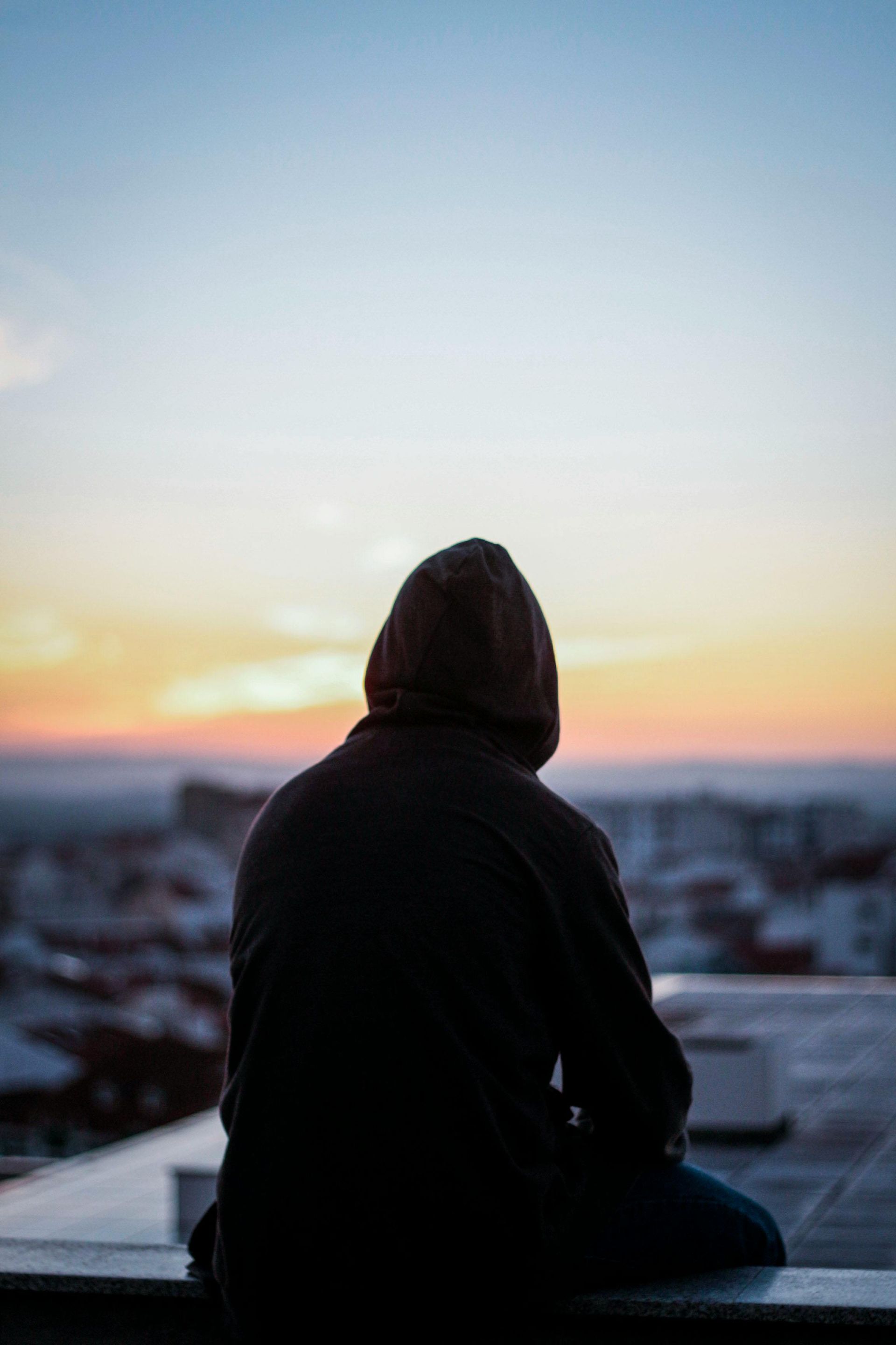 Un homme en sweat à capuche est assis sur un rebord et regarde le coucher du soleil.
